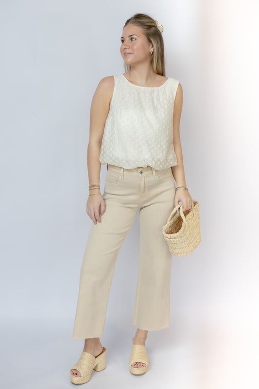 Woman wearing a white sleeveless top and beige pants, holding a woven bag on a plain background