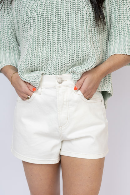 Person wearing a light green knitted sweater and white shorts on a plain background