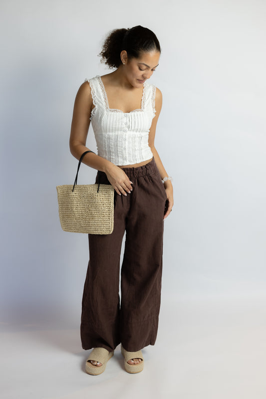 Woman wearing a white top and brown pants holding a straw bag on a plain background