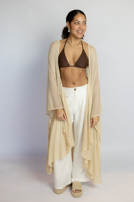 Woman wearing a beige cardigan, brown bikini top, and white pants on a plain background
