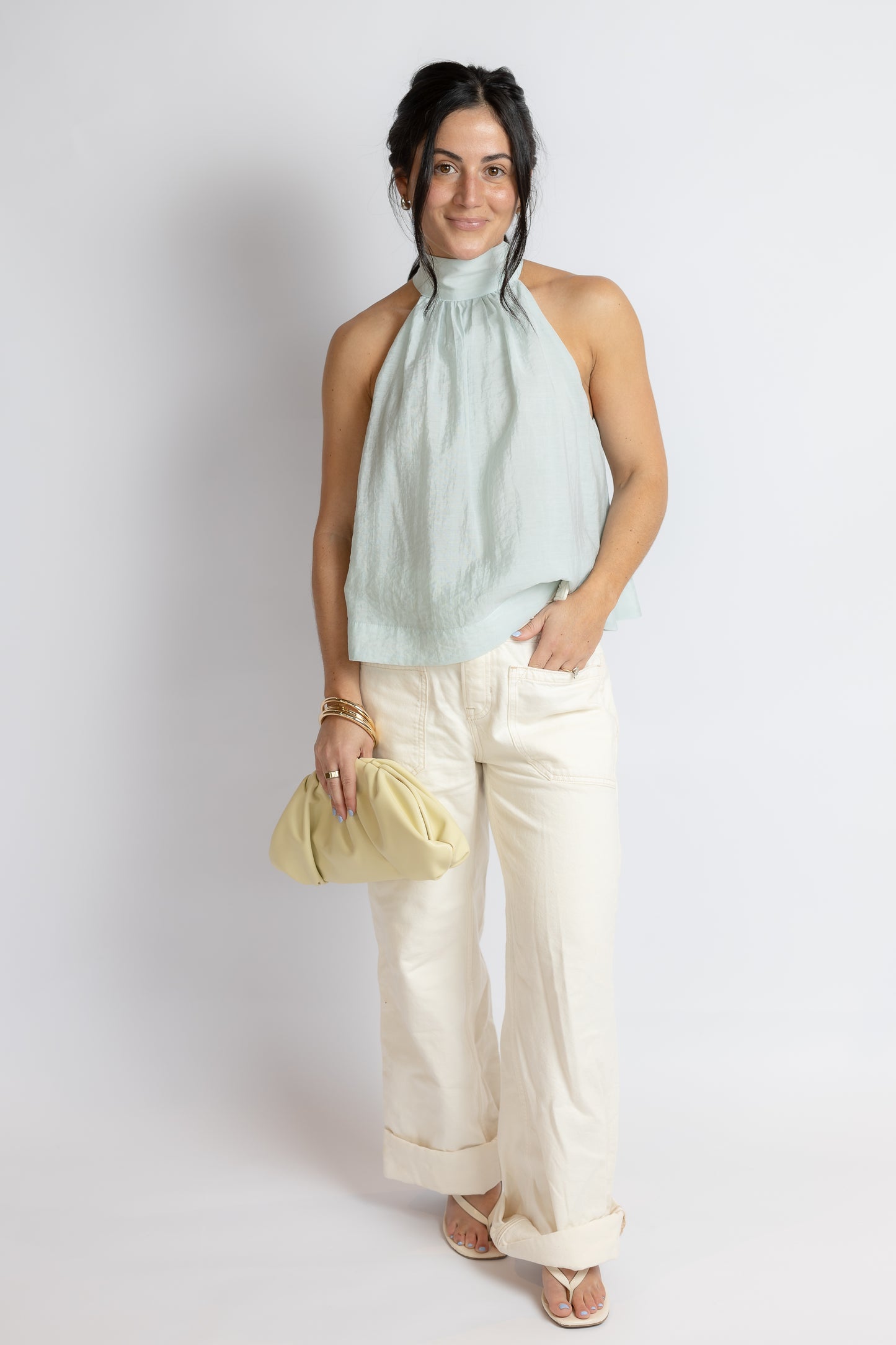 Woman wearing a light green sleeveless top and white pants, holding a yellow clutch against a plain background.