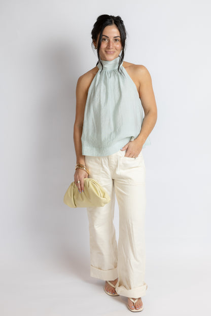 Woman wearing a light green sleeveless top and white pants, holding a yellow clutch against a plain background.