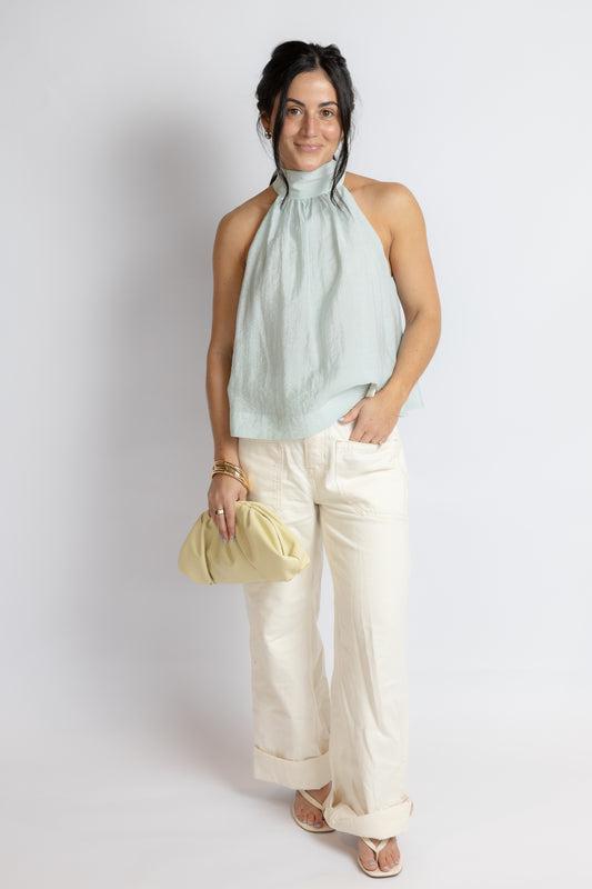 Woman wearing a light green sleeveless top and white pants, holding a yellow clutch against a plain background.
