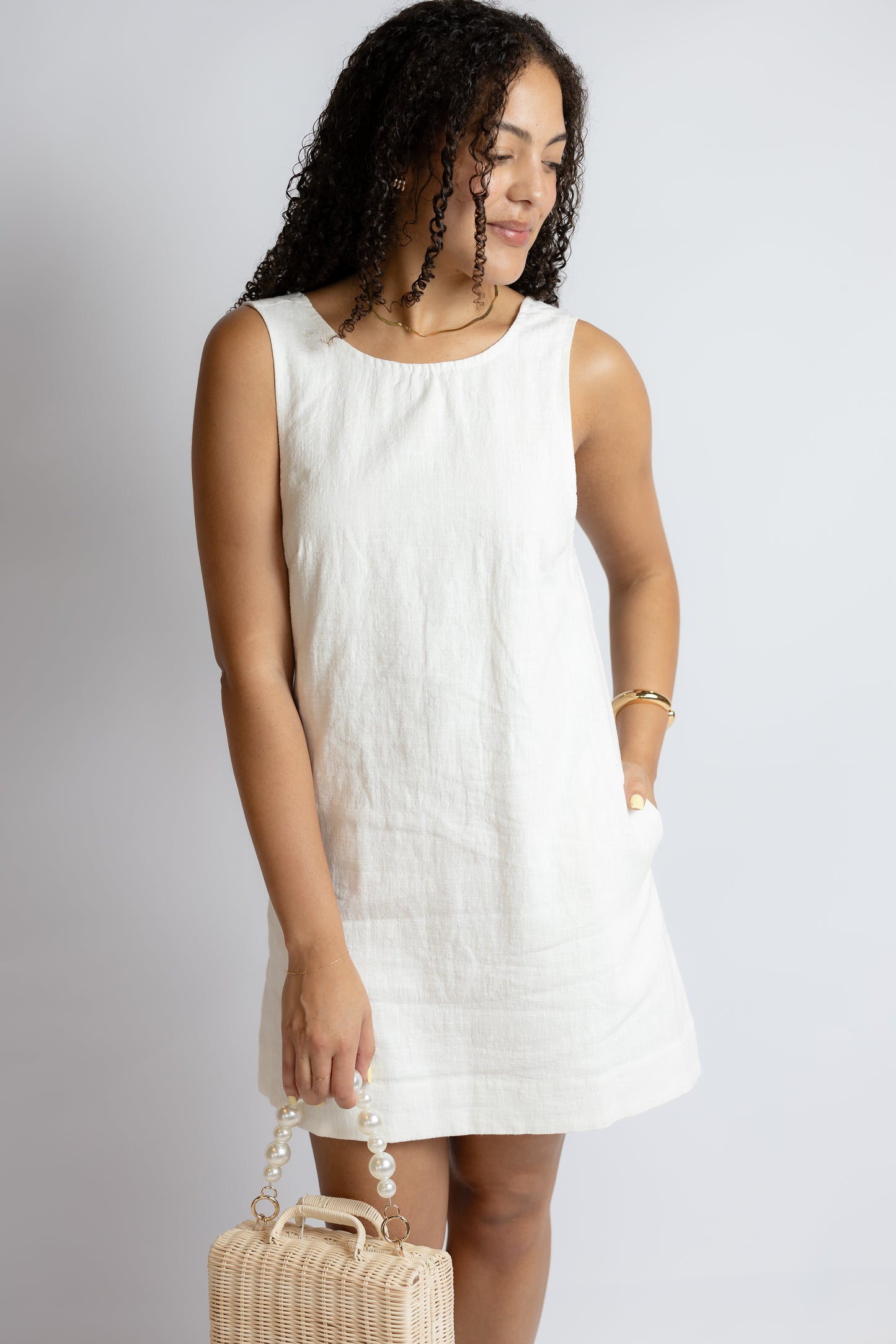 Woman wearing a white sleeveless dress holding a woven handbag against a plain background