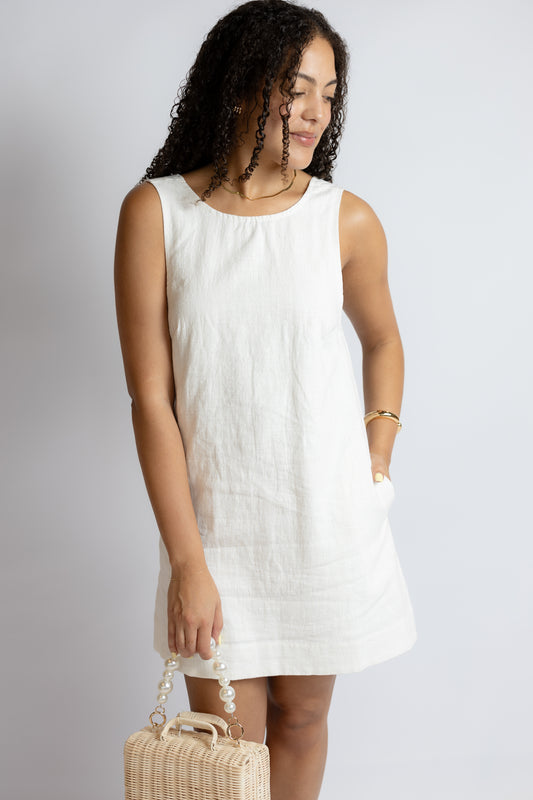 Woman wearing a white sleeveless dress holding a woven handbag against a plain background