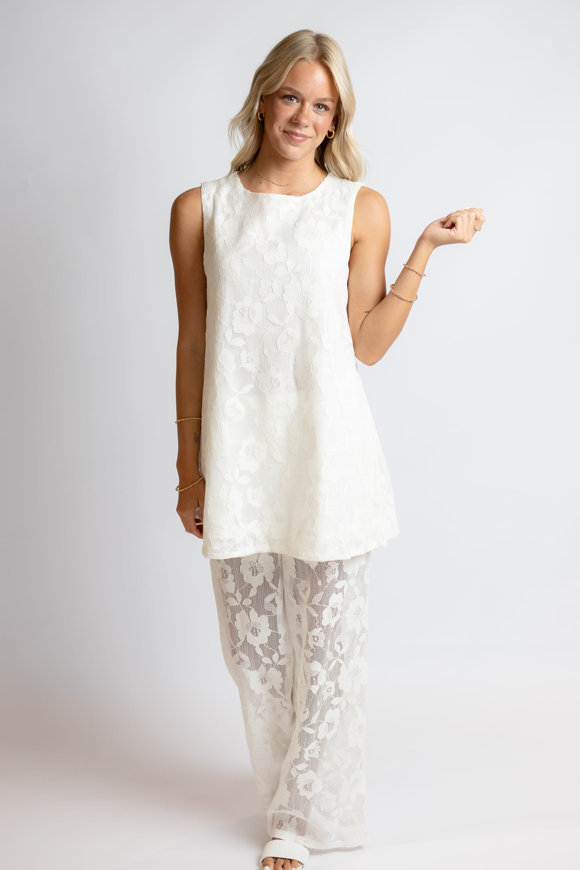Woman wearing a white sleeveless top and lace pants on a plain background