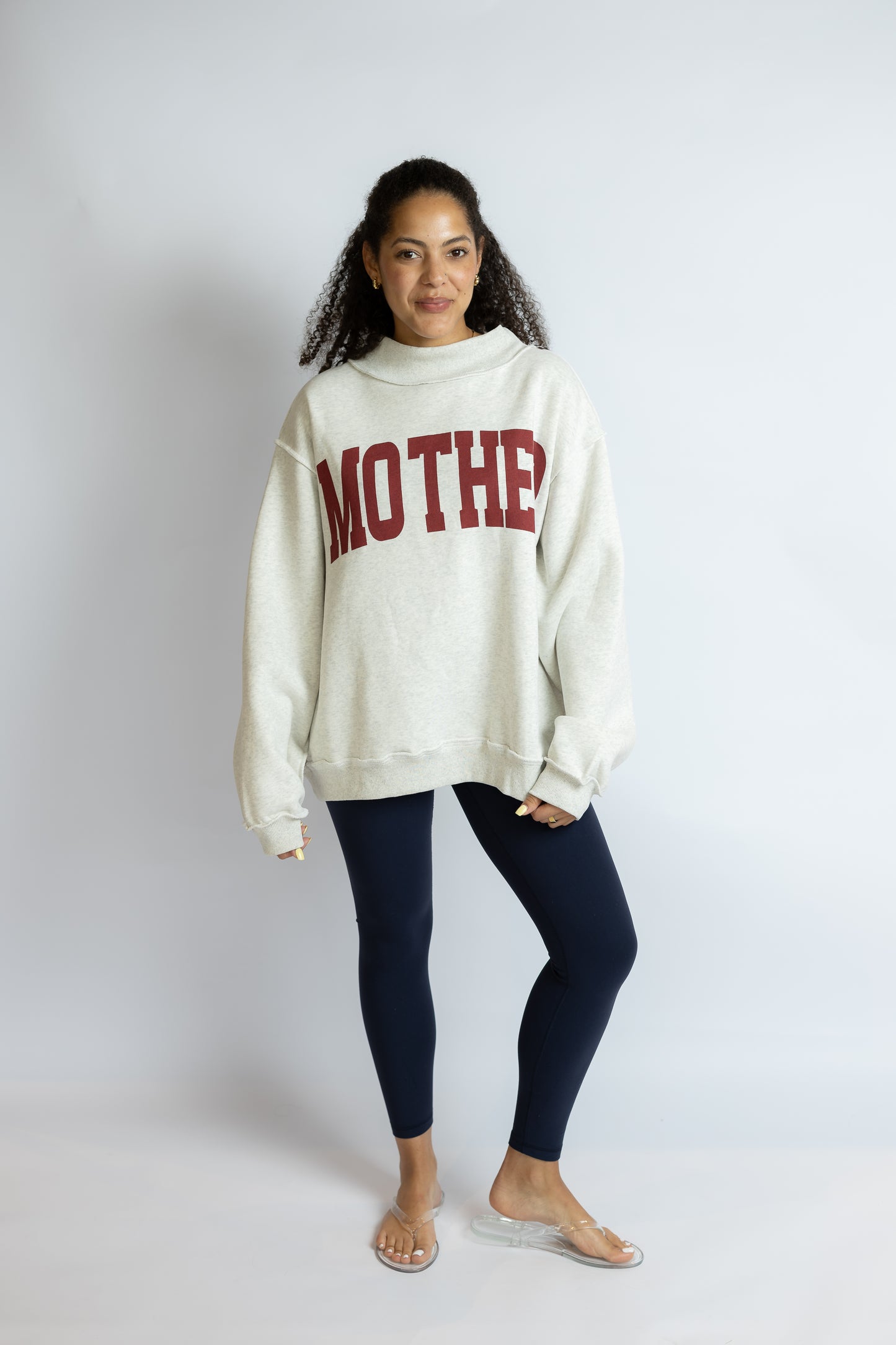 Woman wearing a white sweatshirt with 'MOTHER' printed on it against a plain background