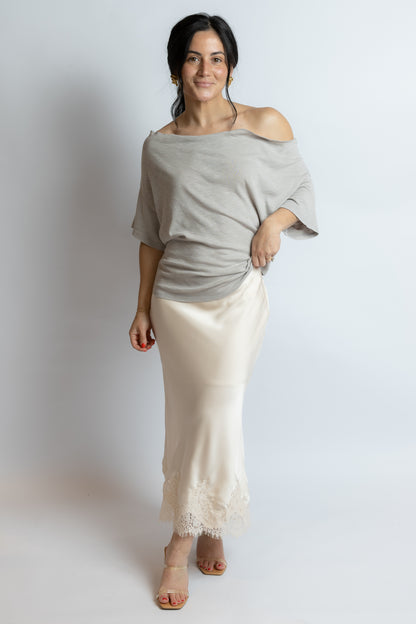 Woman wearing a gray off-shoulder top and white pants against a plain background