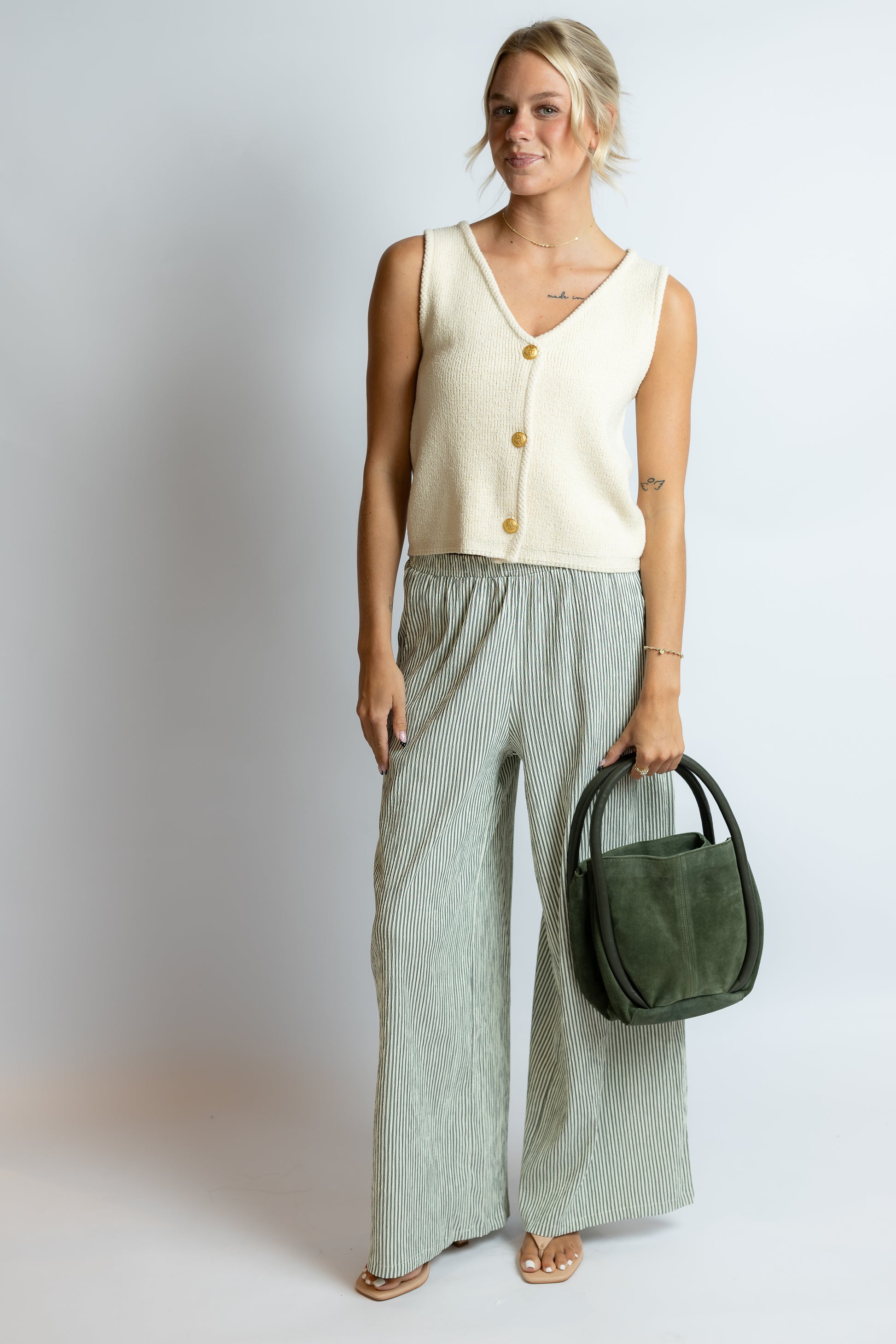 Woman wearing a cream sleeveless top and light green pants, holding a green bag against a white background