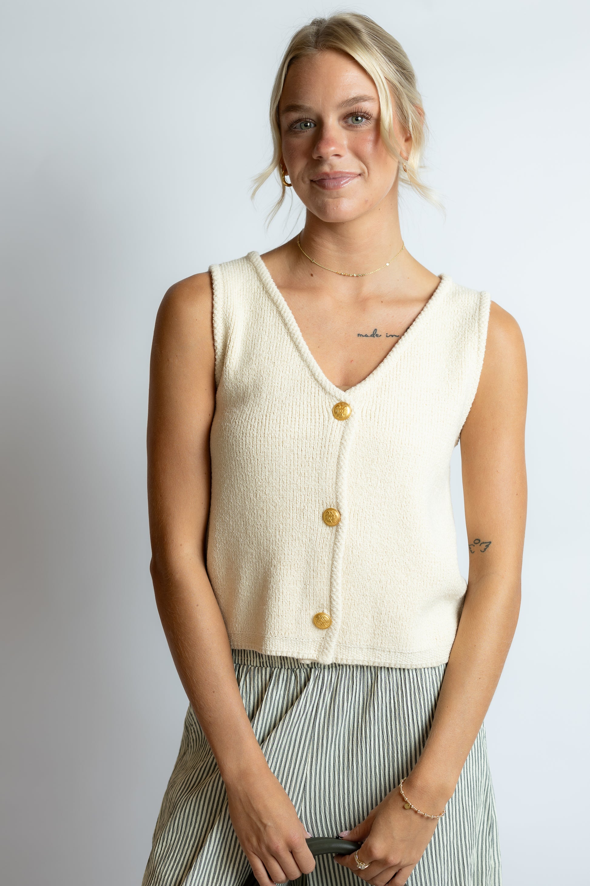 Woman wearing a cream-colored sleeveless top with gold buttons against a plain background