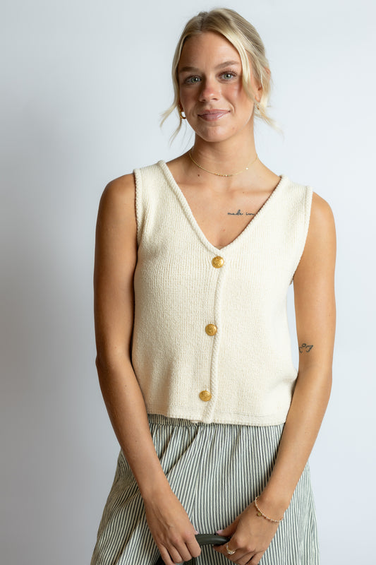Woman wearing a cream-colored sleeveless top with gold buttons against a plain background