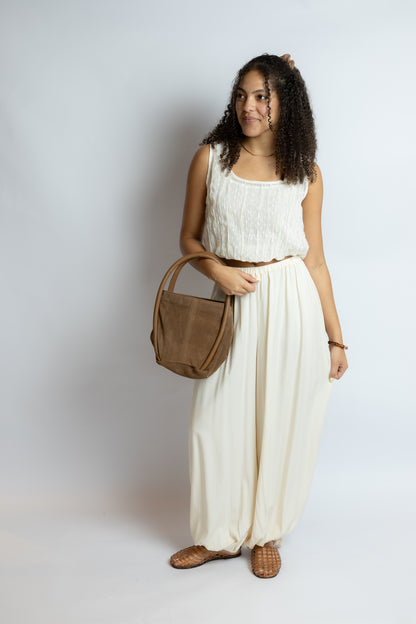 Woman holding a brown handbag wearing a white sleeveless top and cream-colored wide-leg pants on a plain background