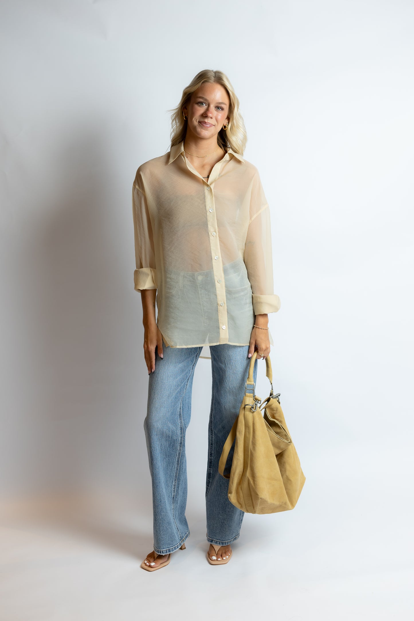 Woman wearing a sheer beige blouse and blue jeans, holding a beige handbag on a white background