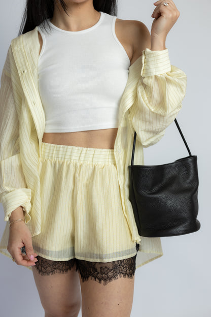 A model wearing a pale yellow striped sheer button-up shirt over a white crop top and matching pale yellow striped shorts with black lace hem, carrying a black bucket bag against a light gray background.