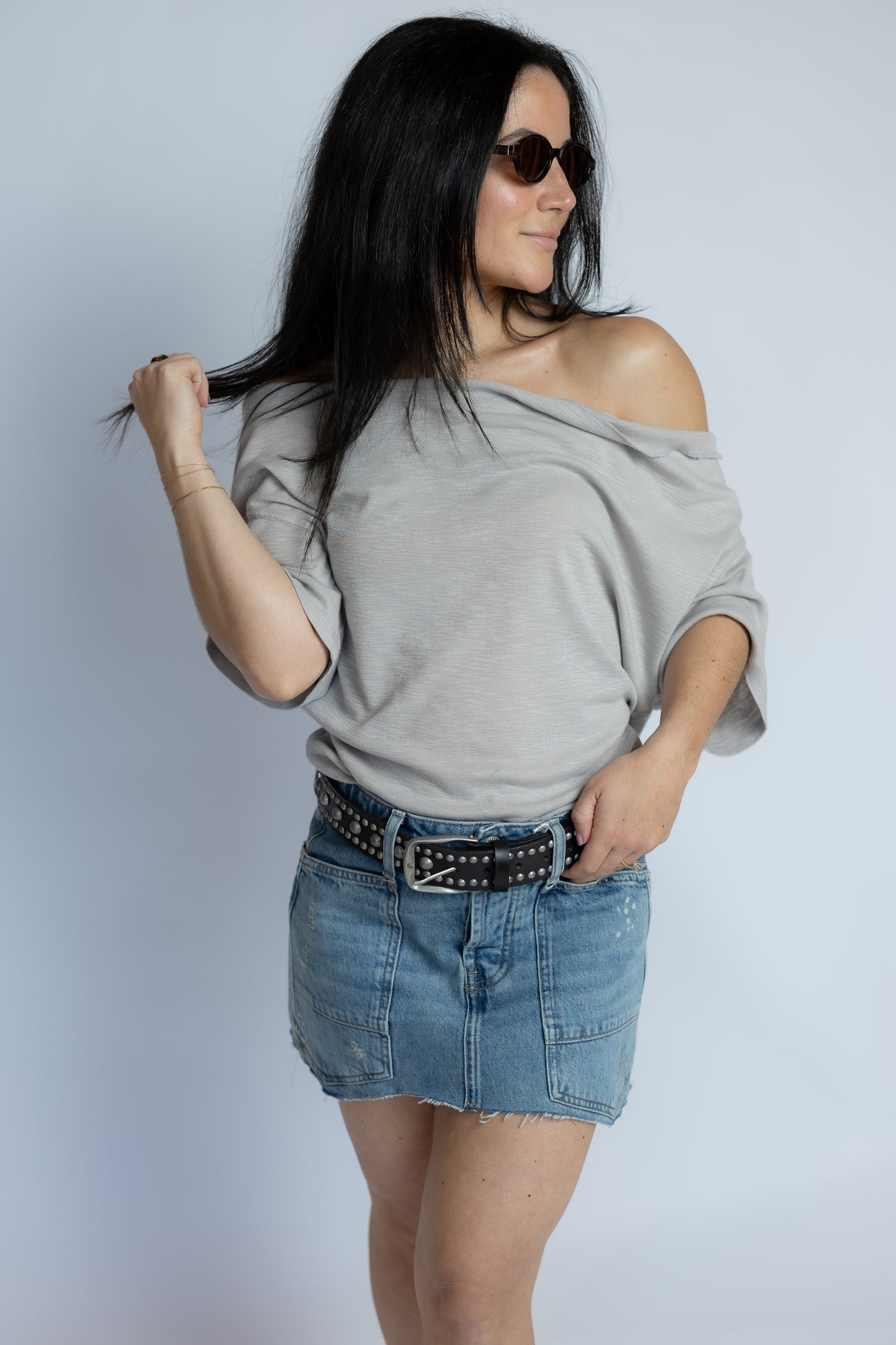 A woman modeling a light gray off-the-shoulder t-shirt and a denim mini skirt with a studded belt, set against a plain light gray background.
