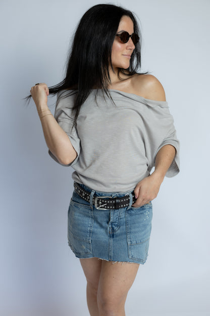 A woman modeling a light gray off-the-shoulder t-shirt and a denim mini skirt with a studded belt, set against a plain light gray background.