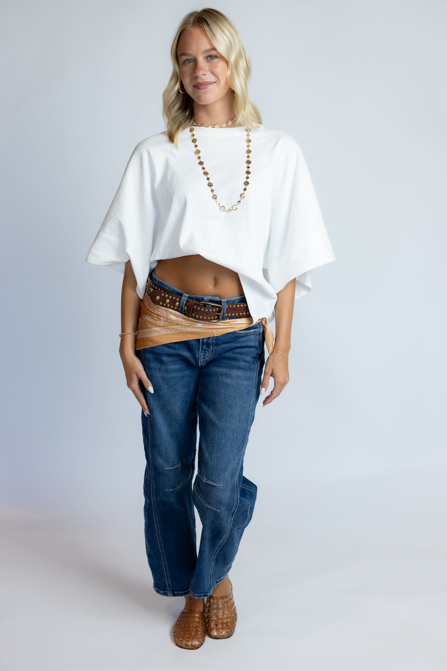 A woman modeling a white oversized short-sleeve top with blue jeans, a brown studded belt, and brown boots against a light gray studio background.