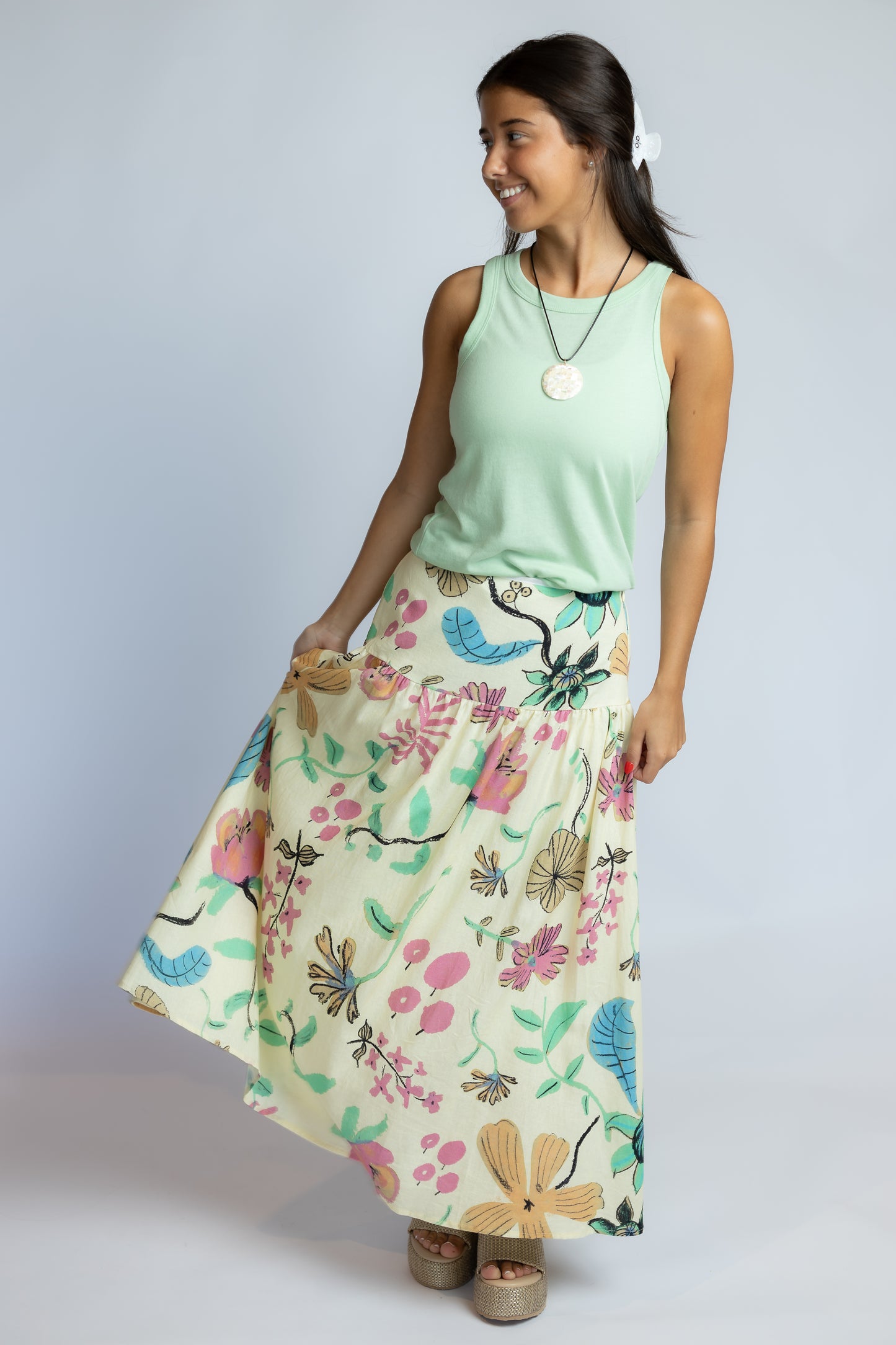 A woman modeling a sleeveless mint green top and a high-waisted, flowing maxi skirt with a colorful floral and butterfly print on a light cream background, set against a plain light gray studio background.