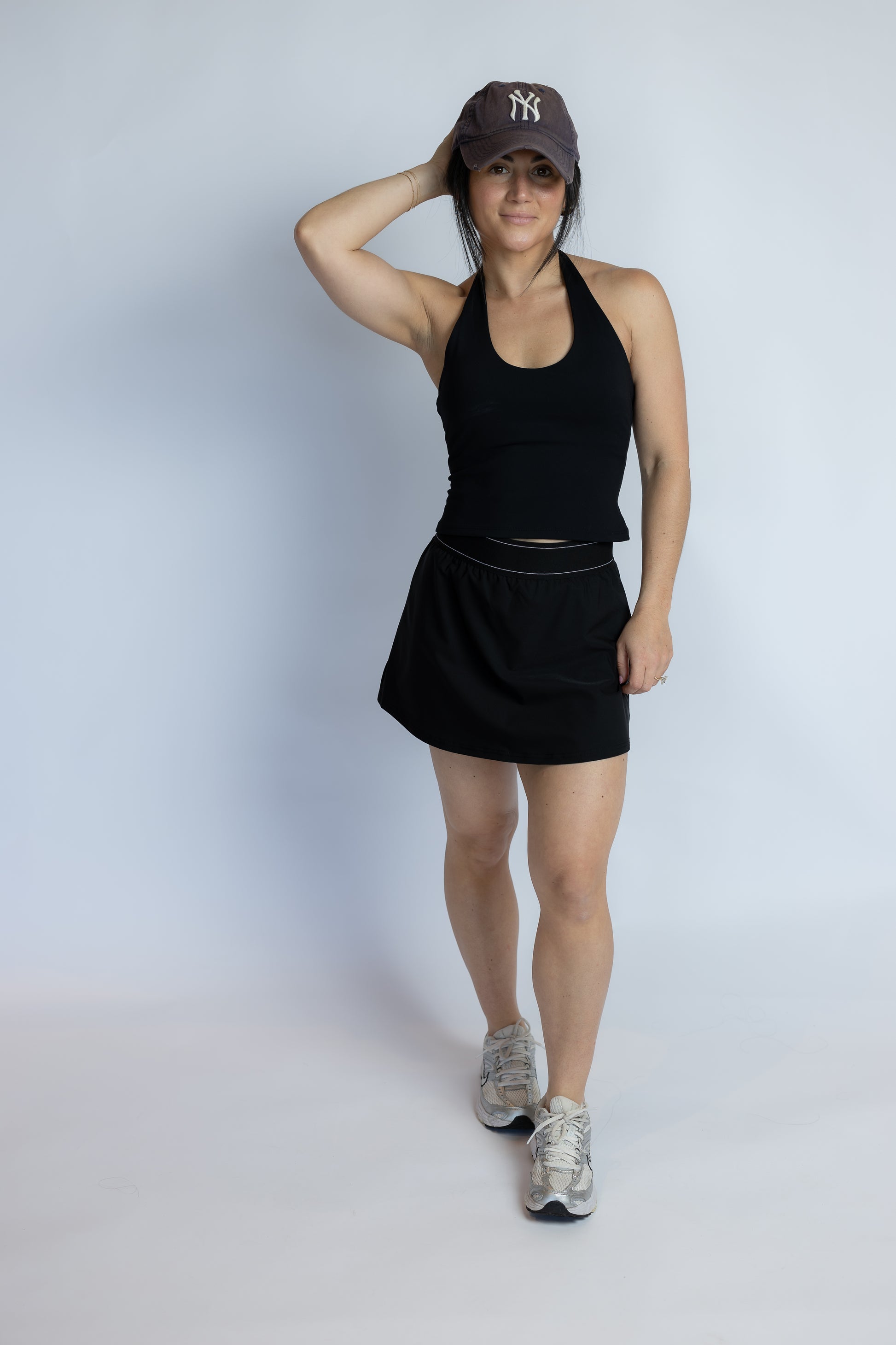A woman modeling a black athletic tank top and matching black tennis skirt, paired with a dark baseball cap and gray running shoes, against a light gray studio background.