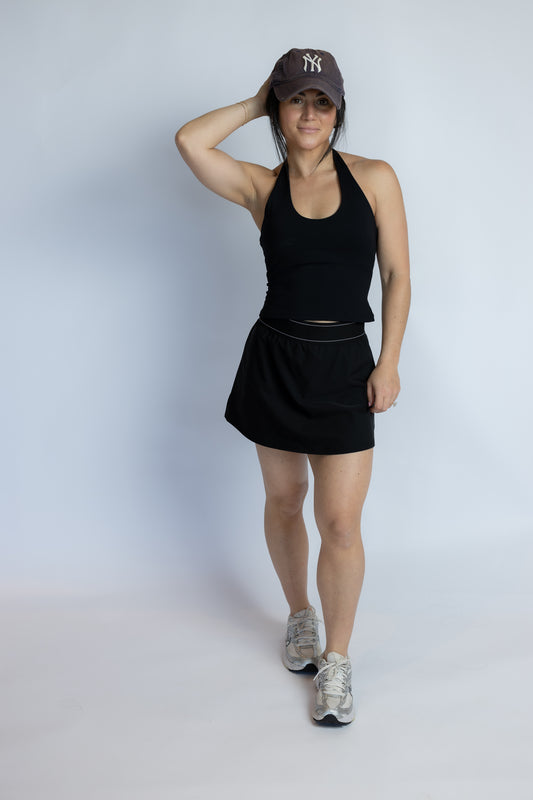 A woman modeling a black athletic tank top and matching black tennis skirt, paired with a dark baseball cap and gray running shoes, against a light gray studio background.