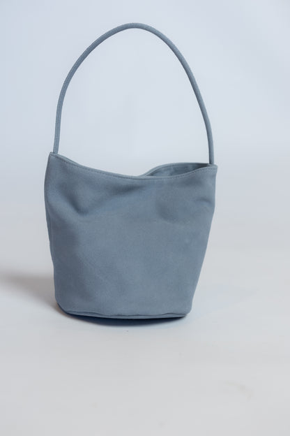 A dusty blue suede bucket bag with a single top handle, photographed against a neutral light gray background.