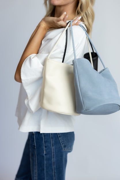 A woman wearing a white blouse and blue jeans carries two large leather tote bags, one in cream and one in dusty blue, against a plain light gray background.