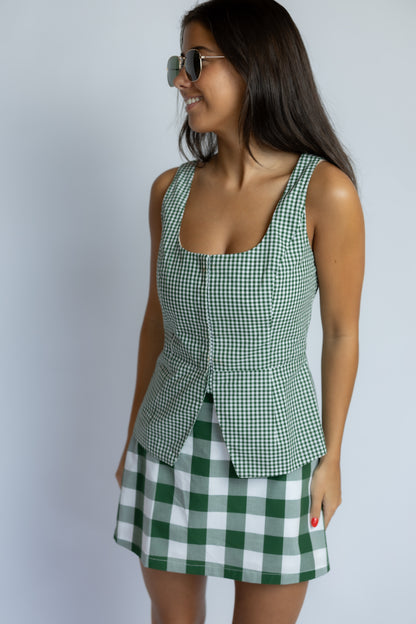 A woman wearing a green and white gingham check peplum top and matching mini skirt, styled with sunglasses, posing against a light gray studio background.