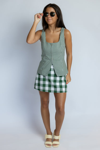 A woman modeling a green and white gingham sleeveless top and matching mini skirt with white heeled sandals against a light gray studio background.