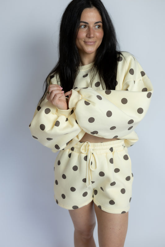 A woman modeling a cream-colored two-piece loungewear set with dark brown polka dots, consisting of a cropped sweatshirt with balloon sleeves and matching shorts, against a plain light gray background.