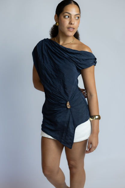 A woman modeling a navy blue off-the-shoulder top with a gathered, ruched design and a gold button detail at the waist, paired with white shorts against a light gray studio background.