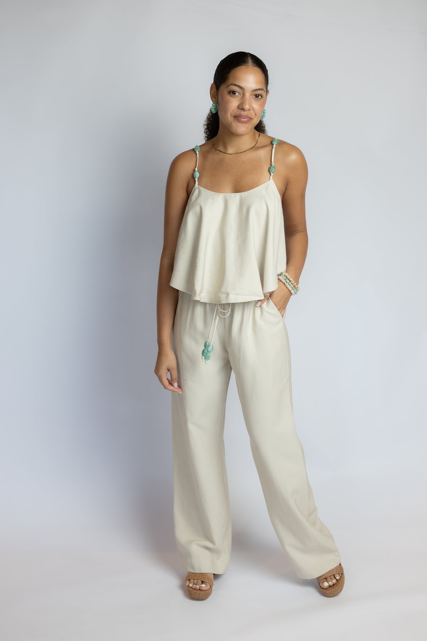 A woman modeling a light cream two-piece lounge set consisting of a sleeveless camisole top and matching wide-leg pants, set against a plain light gray studio background.
