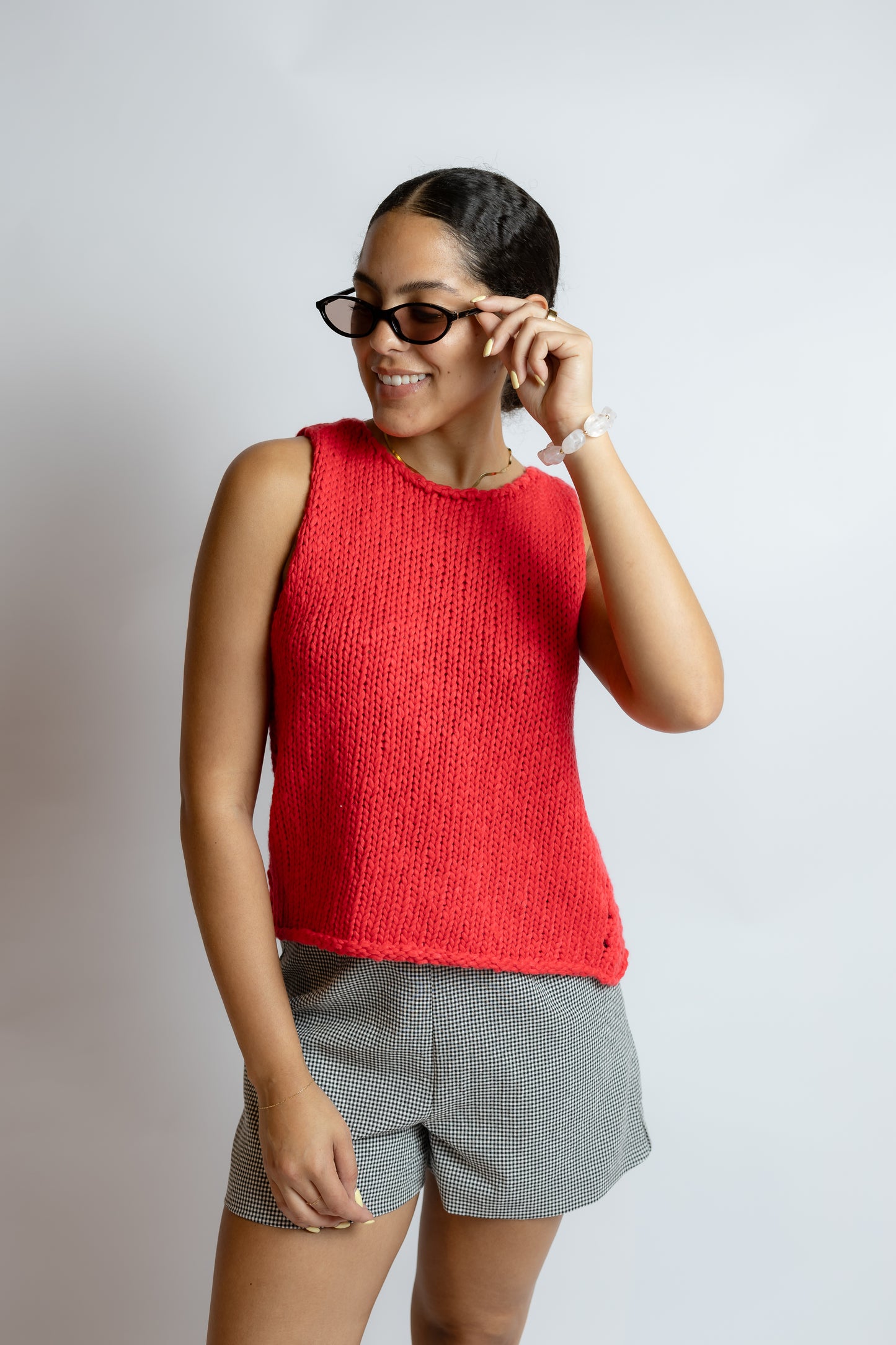 Woman wearing a red sleeveless top and gray shorts, smiling and adjusting sunglasses on a plain background
