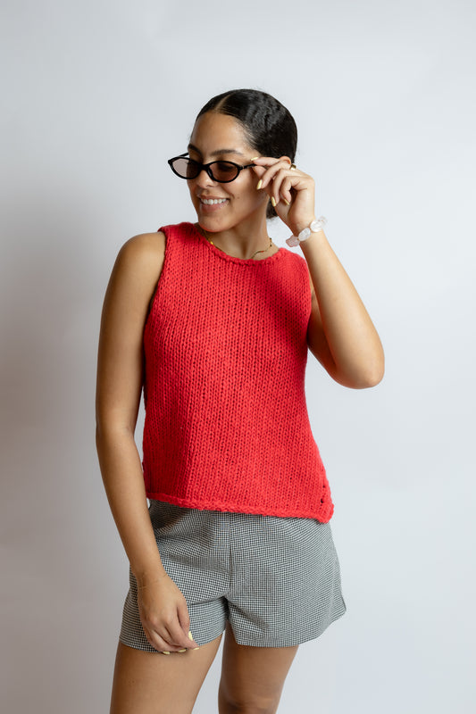 Woman wearing a red sleeveless top and gray shorts, smiling and adjusting sunglasses on a plain background
