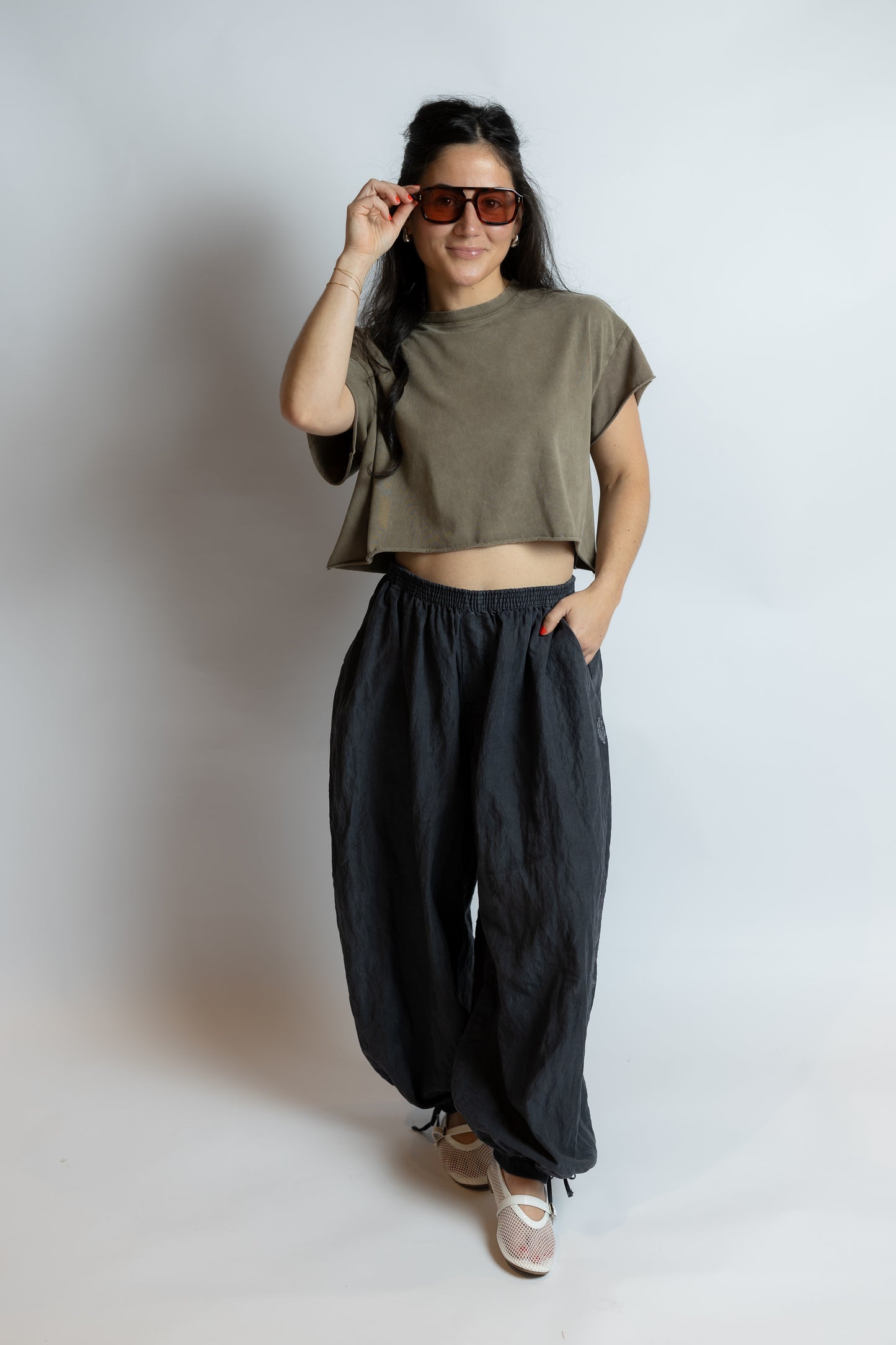 Person wearing a green crop top and black pants, holding sunglasses, on a plain background