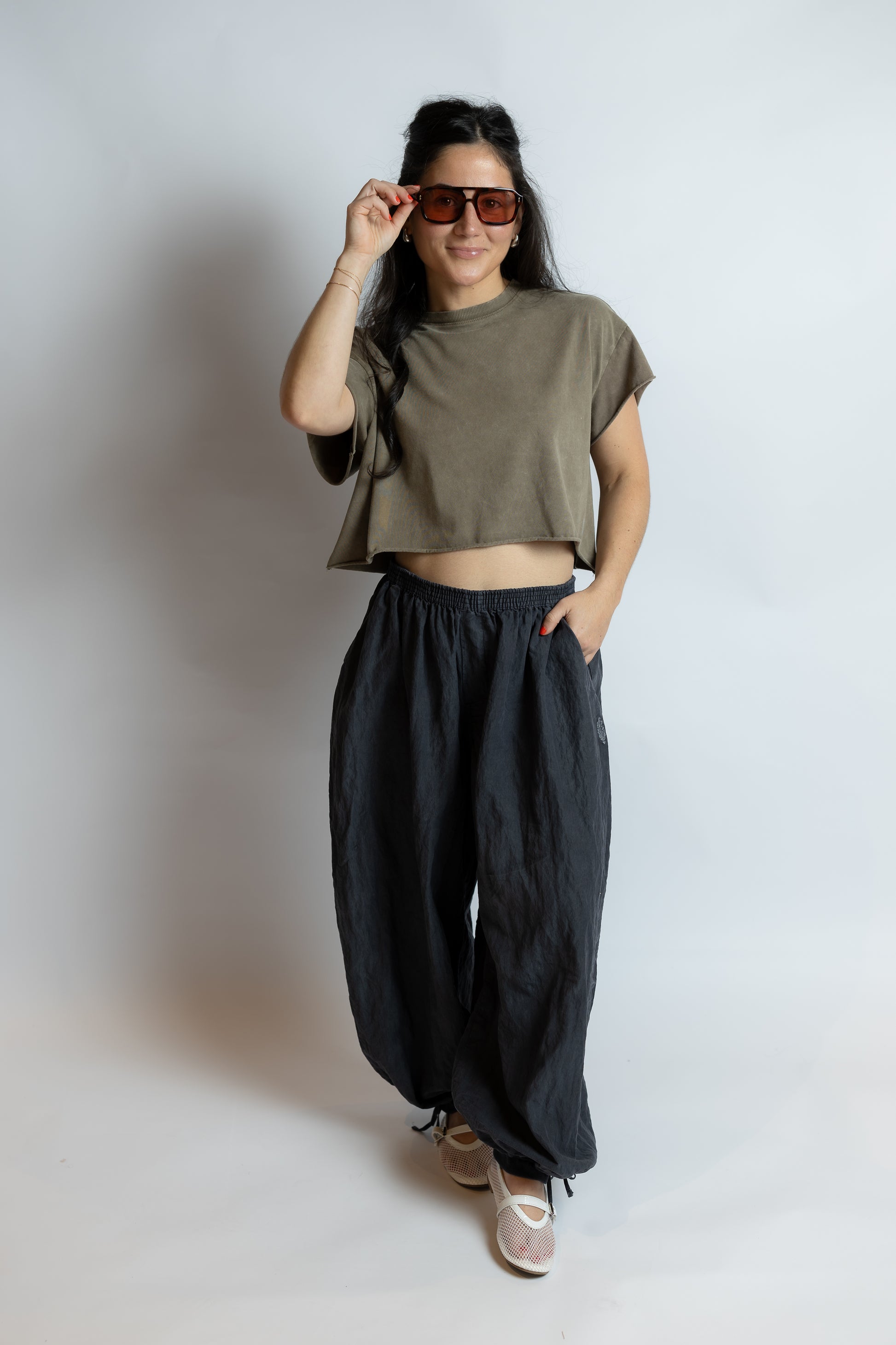 Person wearing a green crop top and black pants, holding sunglasses, on a plain background
