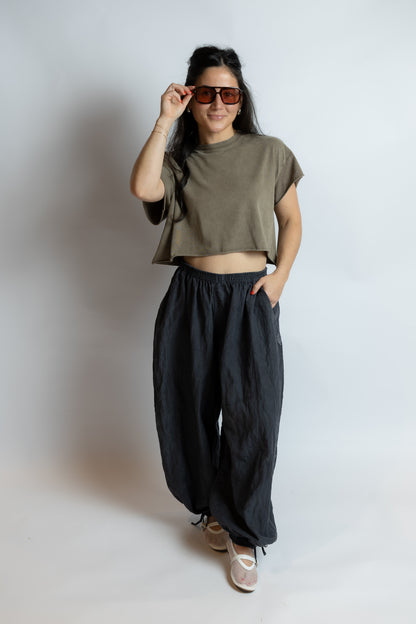 Person wearing a green crop top and black pants, holding sunglasses, on a plain background