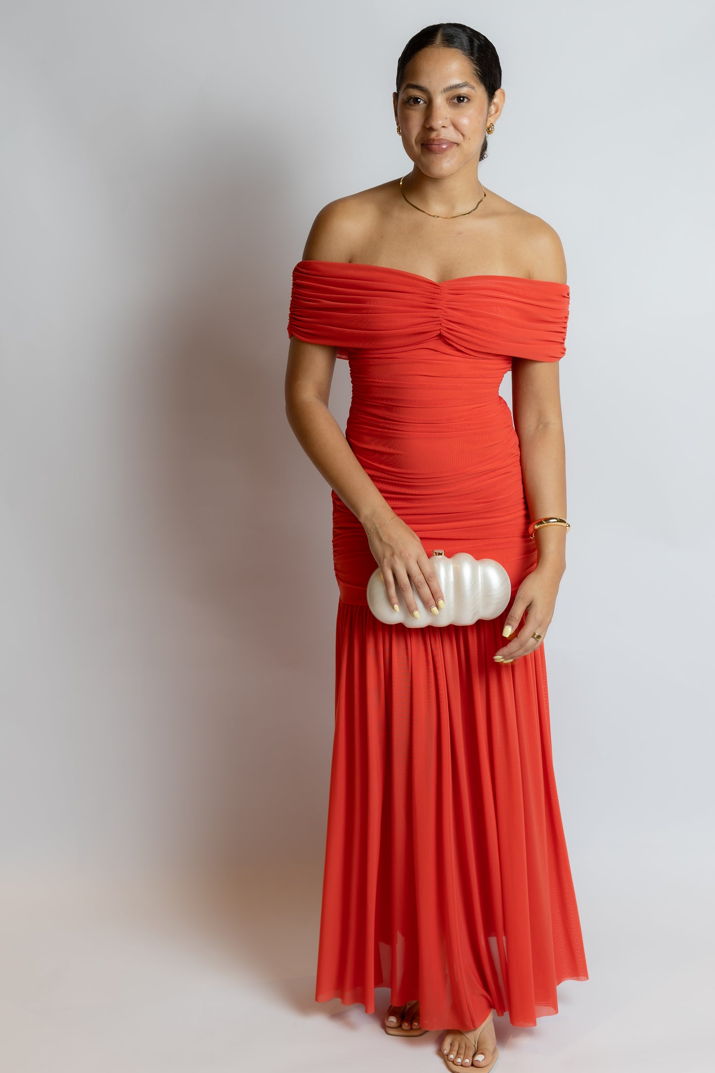 Woman wearing a red off-shoulder dress holding a white clutch against a plain background