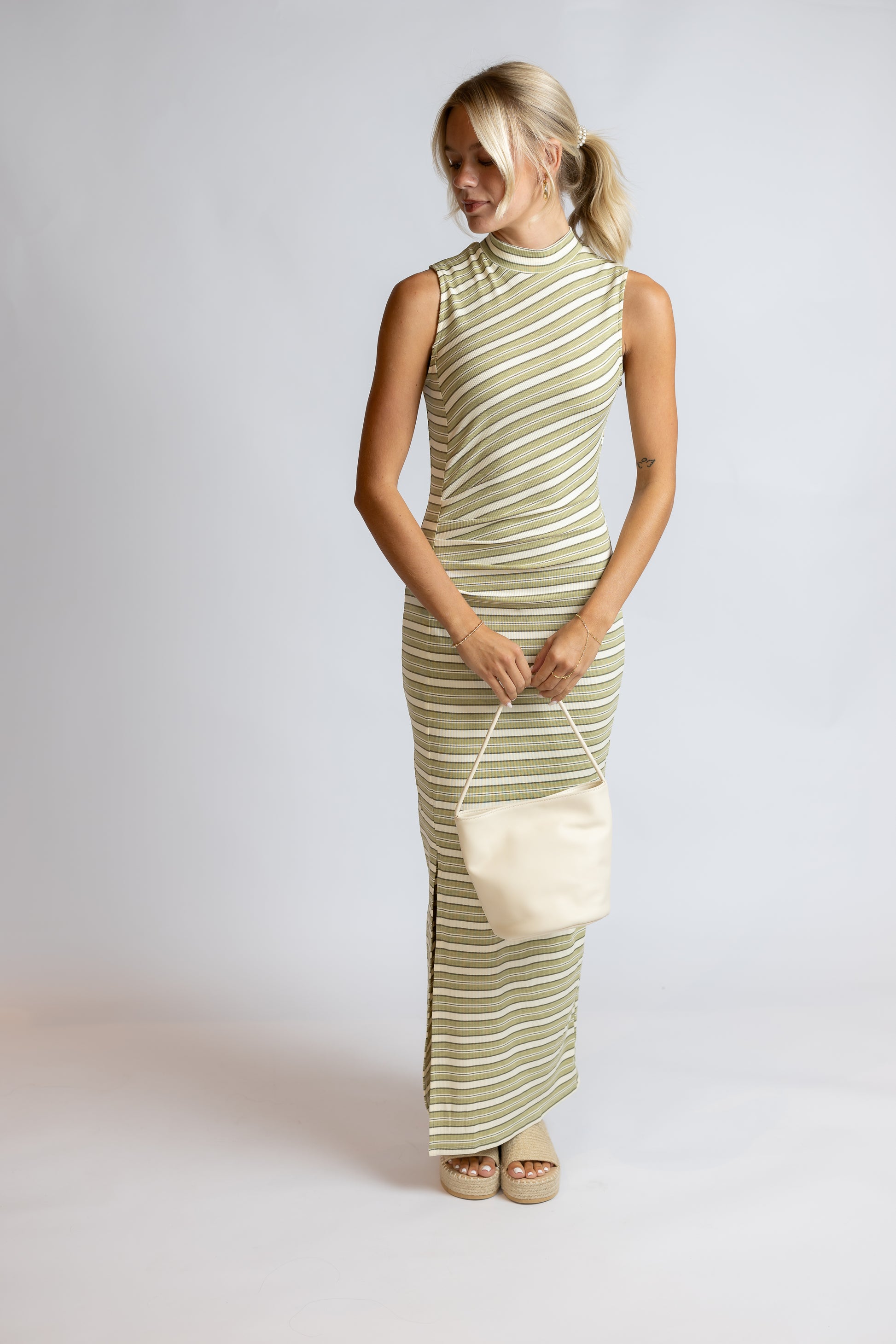 A woman modeling a sleeveless, high-neck maxi dress with diagonal olive green and cream stripes, holding a matching cream handbag against a light gray studio background.