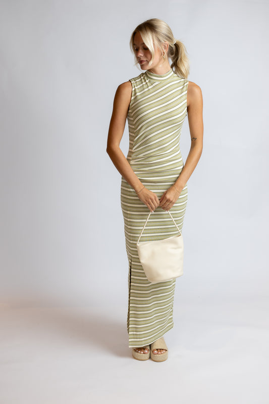 A woman modeling a sleeveless, high-neck maxi dress with diagonal olive green and cream stripes, holding a matching cream handbag against a light gray studio background.