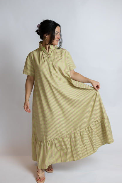 A woman modeling a short-sleeved, high-neck maxi dress with vertical stripes in a muted sage green and cream color, set against a plain light gray studio background.