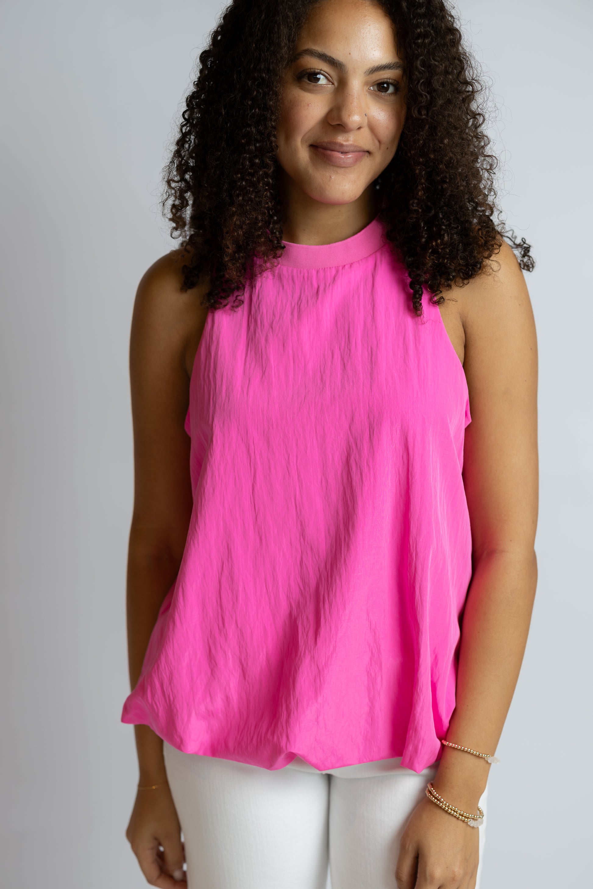 A woman with curly dark hair wearing a bright pink sleeveless mock-neck top and white pants, standing against a light gray studio background.