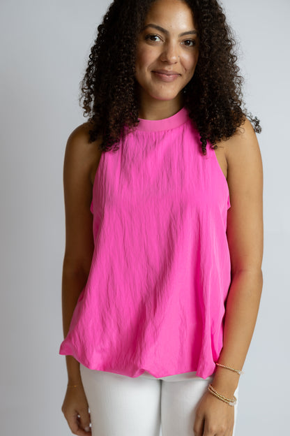 A woman with curly dark hair wearing a bright pink sleeveless mock-neck top and white pants, standing against a light gray studio background.
