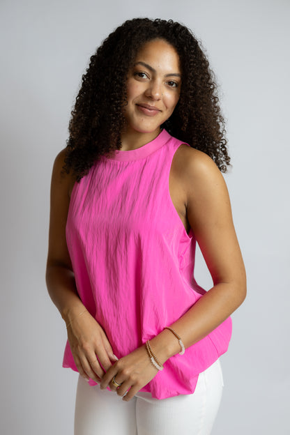 A woman with curly dark hair wearing a sleeveless bright pink top and white pants, standing against a light gray studio background.