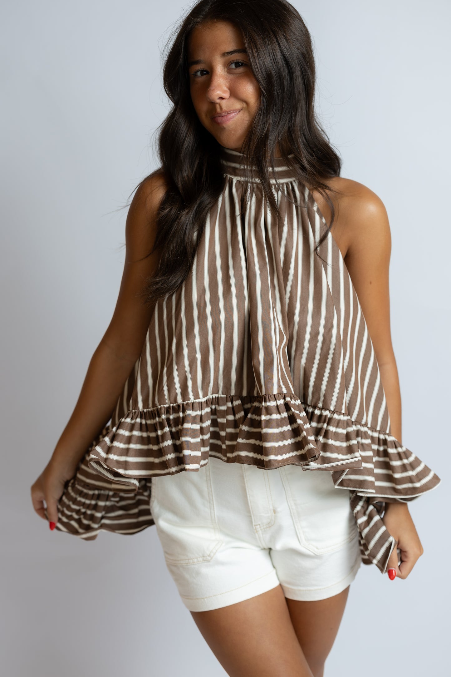 A woman modeling a brown and white vertically striped sleeveless top with a ruffled hem, paired with white shorts, against a light gray studio background.