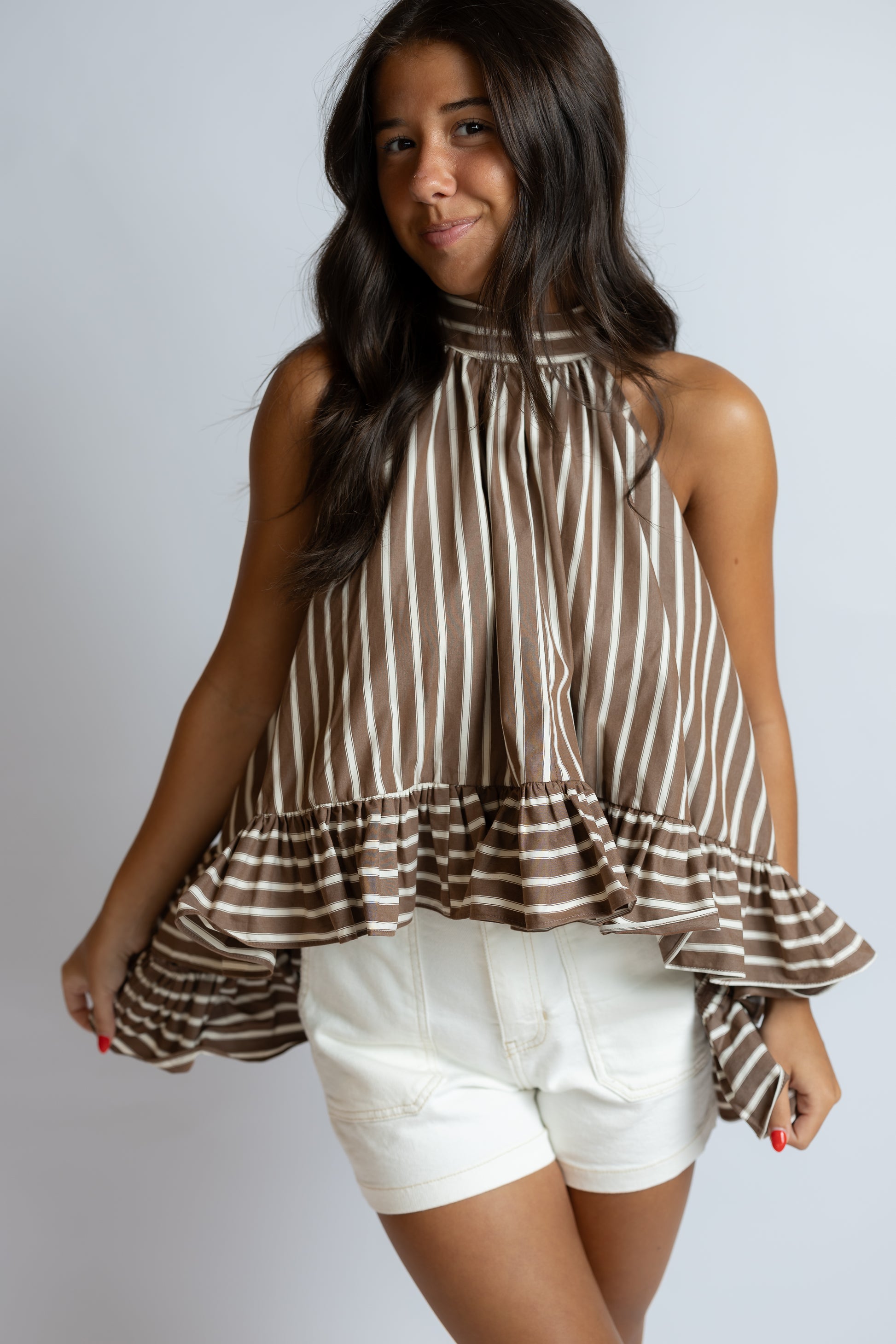 A woman modeling a brown and white vertically striped sleeveless top with a ruffled hem, paired with white shorts, against a light gray studio background.
