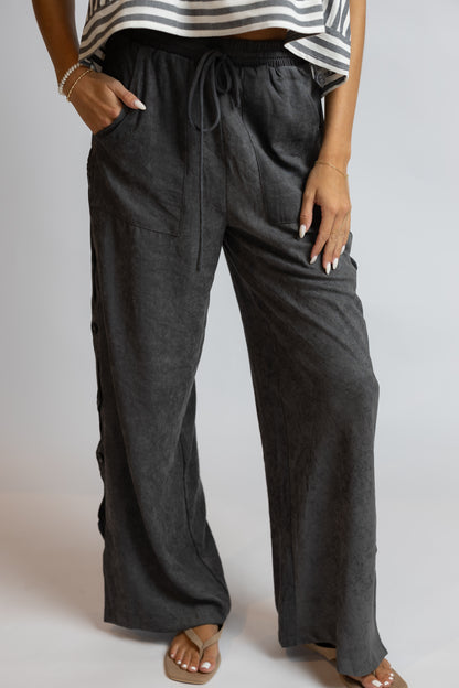 A person wearing dark charcoal gray wide-leg linen pants with a drawstring waist, side pockets, and side slits, paired with a striped top and sandals.