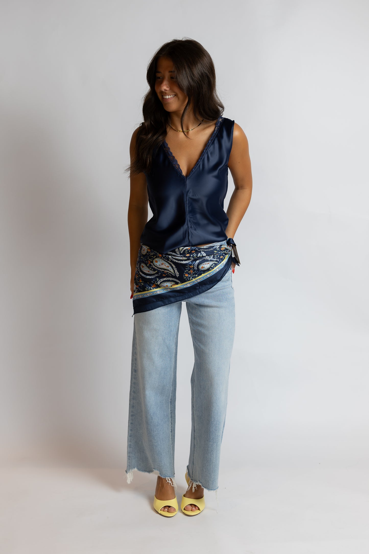 A woman modeling a navy blue satin sleeveless top with a paisley-patterned hem, paired with light-wash wide-leg jeans and yellow sandals, against a plain light gray studio background.