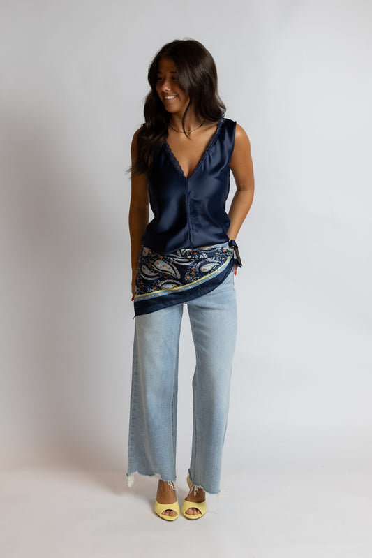 A woman modeling a navy blue satin sleeveless top with a paisley-patterned hem, paired with light-wash wide-leg jeans and yellow sandals, against a plain light gray studio background.