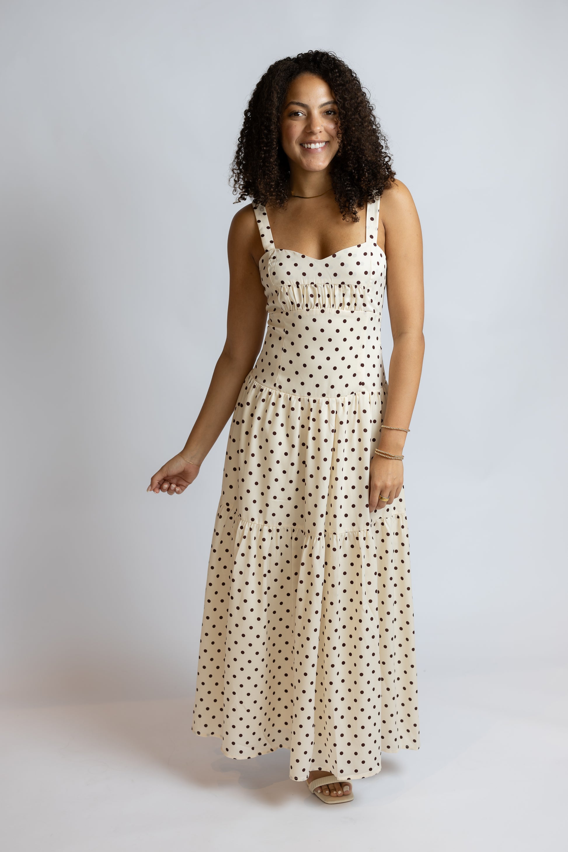 A woman modeling a sleeveless cream maxi dress with a black polka dot pattern against a light gray studio background.