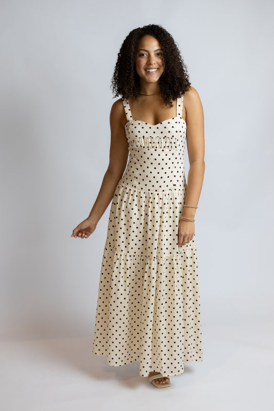 A woman modeling a sleeveless cream maxi dress with a black polka dot pattern against a light gray studio background.
